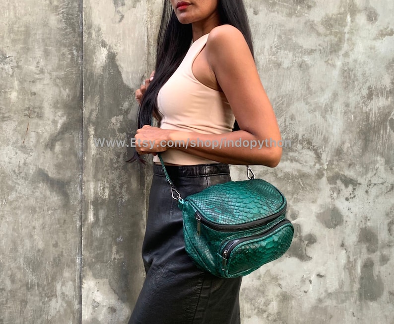 May include: A teal green snakeskin crossbody bag with a zip closure and multiple compartments. The bag is being worn by a person wearing a beige tank top and a black skirt.