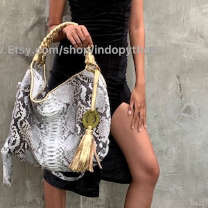 May include: A large, white and grey snakeskin handbag with a gold tassel and a gold circular charm. The bag has a braided gold strap.