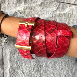 Può includere: Una cintura in pelle di serpente rossa con una fibbia dorata. La cintura è avvolta attorno al braccio di una persona.