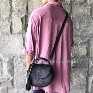 May include: A black leather handbag with a snakeskin pattern and a tassel. The bag has a top handle and a long strap. The bag is being worn by a person wearing a pink shirt.