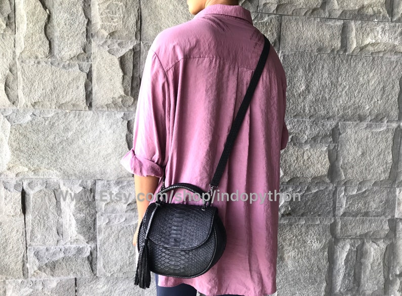 May include: A black leather handbag with a snakeskin pattern and a tassel. The bag has a top handle and a long strap. The bag is being worn by a person wearing a pink shirt.