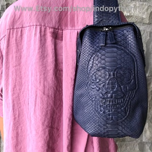 May include: A navy blue sling bag featuring a skull design embossed on the front. The bag is made from a textured material, possibly leather. It has a zip closure and a single strap. The bag is worn over the shoulder.