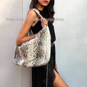 Pode incluir: Uma bolsa tote grande de pele de cobra branca e marrom com uma alça de corrente prateada. A bolsa está sendo segurada por uma pessoa que está vestindo um vestido preto.