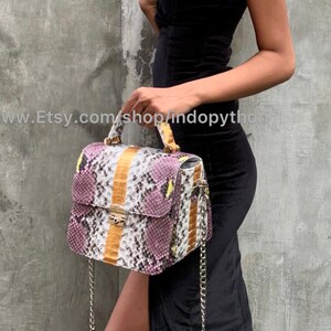 May include: A small, square handbag made of purple, white, and yellow snakeskin. The bag has a top handle and a chain strap. The bag is being held by a person wearing a black dress.