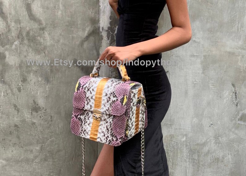 May include: A small, square handbag made of purple, white, and yellow snakeskin. The bag has a top handle and a chain strap. The bag is being held by a person wearing a black dress.