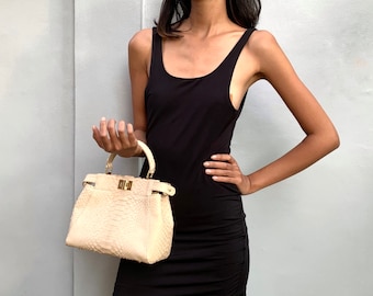 May include: A woman wearing a black sleeveless dress holds a small, light beige handbag with a gold clasp. The handbag has a textured, snakeskin-like pattern.