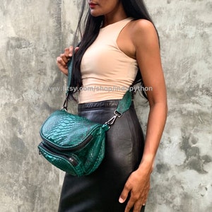 May include: A green snakeskin crossbody bag with a black strap and a zip closure. The bag is being worn by a person wearing a light pink tank top and a black skirt.