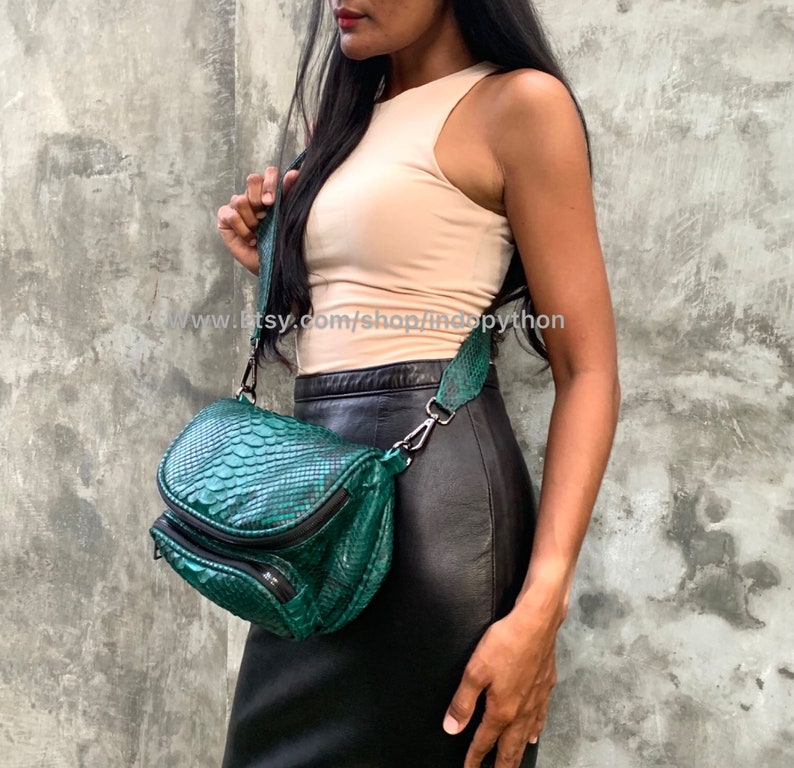 May include: A green snakeskin crossbody bag with a black strap and a zip closure. The bag is being worn by a person wearing a light pink tank top and a black skirt.