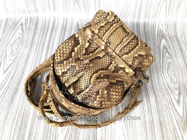 May include: A tan and brown snakeskin crossbody bag with a flap closure and adjustable strap. The bag features a textured pattern and a dark zipper. The strap is made of the same snakeskin material. The bag is designed to be worn over the shoulder.