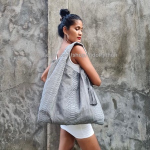 Pode incluir: Uma bolsa tote de couro de pitão cinza com um tassel. A bolsa tem uma superfície texturizada e escamosa e é grande o suficiente para carregar os itens essenciais do dia a dia.