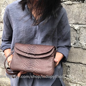 May include: A brown snakeskin clutch bag with a flap closure and a long strap. The bag is being held by a person wearing a grey shirt.