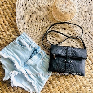 May include: A black leather crossbody bag with a snakeskin pattern. The bag is on a woven mat with a straw hat and denim shorts.