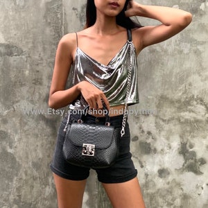 May include: A silver metallic top with a cowl neck and a chain strap. The top is paired with black denim shorts and a black snakeskin handbag with a silver chain strap.