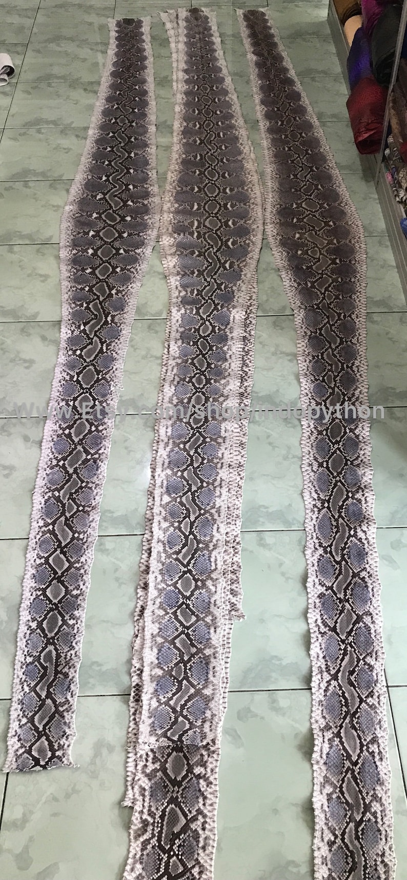 May include: Three long strips of light gray python skin with a dark gray pattern. The skin is laid out flat on a tiled floor.