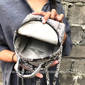 K&ouml;nnte beinhalten: Ein kleiner, grau-schwarzer Schlangenleder-Rucksack mit Rei&szlig;verschluss und einem oberen Griff. Der Rucksack ist offen und das Innere ist mit einem hellgrauen Stoff ausgekleidet.