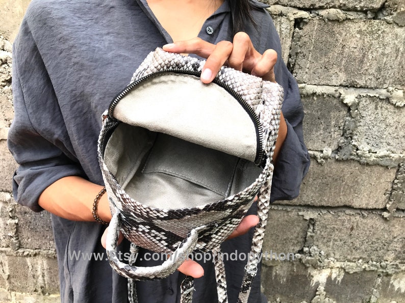 K&ouml;nnte beinhalten: Ein kleiner, grau-schwarzer Schlangenleder-Rucksack mit Rei&szlig;verschluss und einem oberen Griff. Der Rucksack ist offen und das Innere ist mit einem hellgrauen Stoff ausgekleidet.