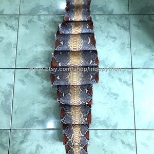 May include: A stack of python skin pieces, displaying a natural pattern of grey, tan, and reddish-brown hues. The arrangement highlights the unique textures and colours of the reptile skin, with the Etsy shop URL visible at the bottom.