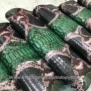 Pode incluir: Close-up de vários pedaços de couro de pitão. O couro tem um padrão de escamas pretas, verdes e castanhas. As escamas verdes são de uma cor vibrante e sólida, enquanto as outras escamas têm uma aparência manchada. O couro é provavelmente para artesanato.