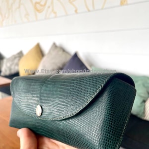 Op de afbeelding: Een donkergroene, getextureerde brillenkoker met een drukknoopsluiting. De koker heeft een reptielachtig patroon en een gebogen flap. De koker wordt in een hand gehouden.