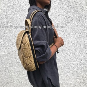 Puede incluir: Una mochila de piel de serpiente de color beige con una cremallera negra y correas negras. La mochila la lleva puesta una persona que lleva una camisa azul oscuro.