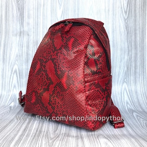 May include: A red snakeskin backpack with black accents and a zipper closure. The backpack has a small pocket on the front and a website address printed on the bottom: Etsy.com/shop/indopython.