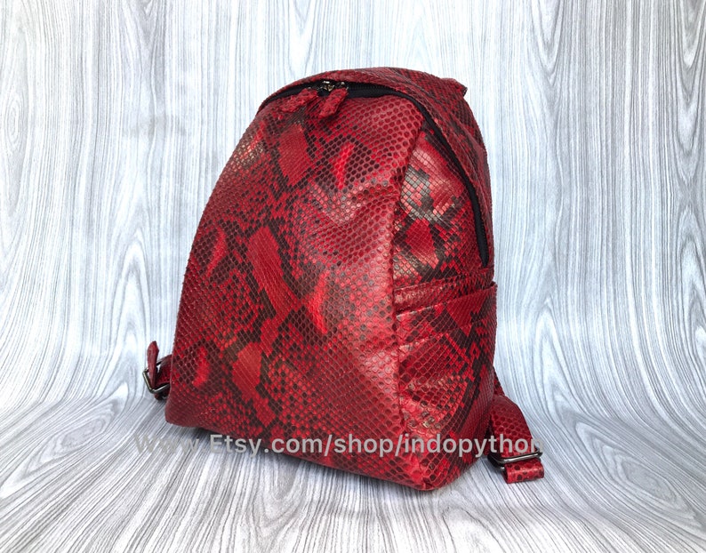 May include: A red snakeskin backpack with black accents and a zipper closure. The backpack has a small pocket on the front and a website address printed on the bottom: Etsy.com/shop/indopython.