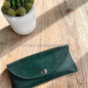 Op de afbeelding: Een donkergroene, getextureerde leren brillenkoker met een drukknoopsluiting. De koker is rechthoekig met een gebogen flap en een zilverkleurige drukknop. Een kleine cactus in pot staat links op een houten oppervlak.