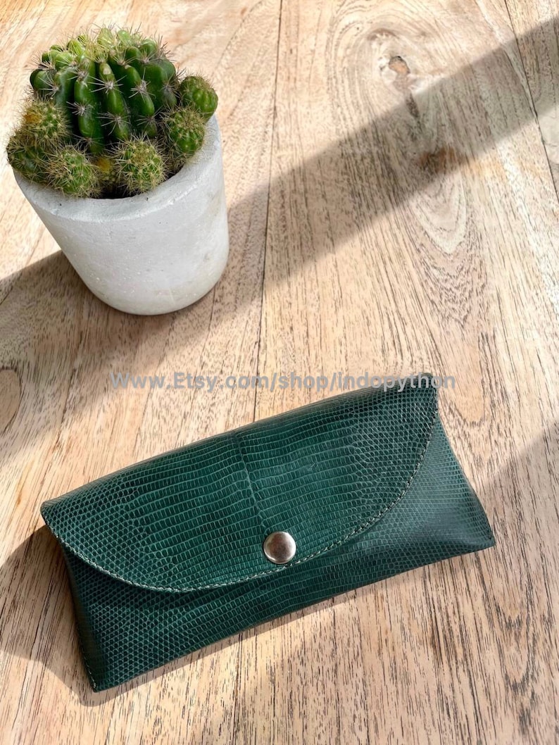 Op de afbeelding: Een donkergroene, getextureerde leren brillenkoker met een drukknoopsluiting. De koker is rechthoekig met een gebogen flap en een zilverkleurige drukknop. Een kleine cactus in pot staat links op een houten oppervlak.