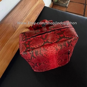 May include: A red and black snakeskin backpack with a zipper closure. The backpack is made of genuine python leather and has a unique, textured design.