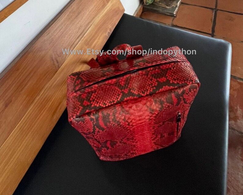 May include: A red and black snakeskin backpack with a zipper closure. The backpack is made of genuine python leather and has a unique, textured design.