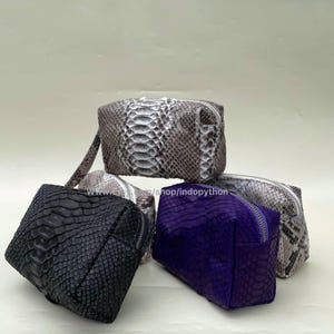 May include: Four different coloured python skin cosmetic bags. The bags are all rectangular and have a zipper closure. The bags are stacked on top of each other. The top bag is a light brown colour with a white pattern. The second bag is a white colour with a brown pattern. The third bag is a black colour with a white pattern. The bottom bag is a purple colour with a white pattern.
