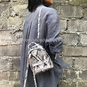 K&ouml;nnte beinhalten: Ein kleiner, wei&szlig;er und schwarzer Schlangenleder-Rucksack mit Rei&szlig;verschluss. Der Rucksack wird von einer Person getragen, die ein graues Hemd tr&auml;gt.