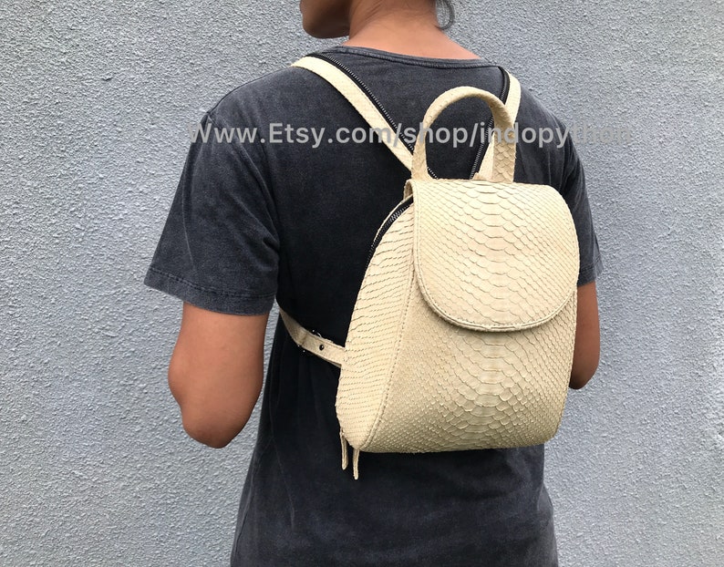 May include: A cream-colored snakeskin backpack with a zipper closure and a top handle. The backpack is being worn by a person.