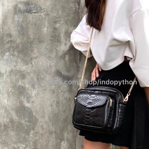 May include: A black snakeskin crossbody bag with a gold chain strap and a front pocket. The bag is worn by a person wearing a white shirt and a black skirt.