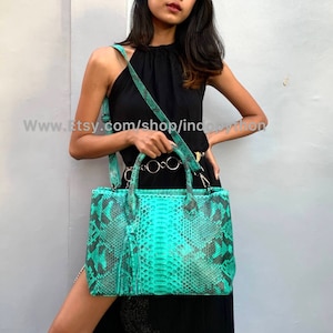 Puede incluir: Un bolso de mano de piel de pitón verde turquesa con asa superior y bandolera. El bolso tiene una forma estructurada y un cierre de solapa frontal.