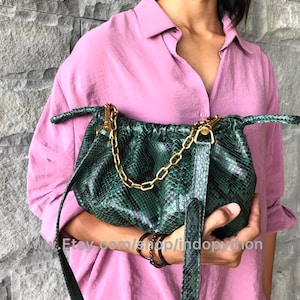 May include: A green snakeskin handbag with a gold chain strap. The bag has a drawstring closure and is held by a person wearing a pink shirt.