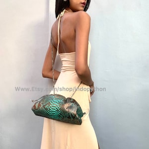 May include: A green and brown snakeskin clutch purse with a drawstring closure. The purse is being worn by a person as a shoulder bag.