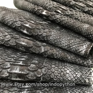 以下が含まれることがあります： 黒と白のパイソンレザーの断片をクローズアップ。レザーは、質感のある鱗状の表面をしています。レザーは折り畳まれ、互いに積み重ねられています。画像の下部に「www.Etsy.com/shop/indopython」というテキストが表示されています。