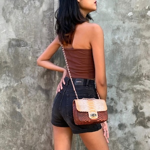 Puede incluir: Un bolso de hombro de piel de serpiente con textura marrón y beige con una correa de cadena dorada. El bolso tiene un cierre de solapa con un broche dorado.