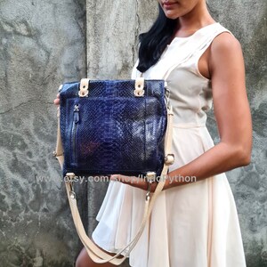May include: A blue python leather tote bag with a beige shoulder strap. The bag has a zippered pocket on the front and silver hardware.