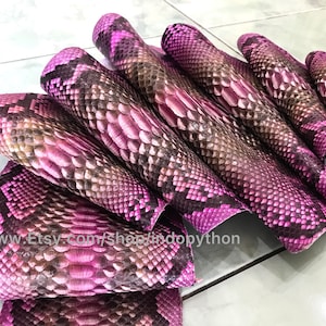 以下が含まれることがあります： ピンクとブラウンのパイソン革の束。革は折り畳まれ、扇形に配置されています。革はテクスチャのあるパターンを持っています。