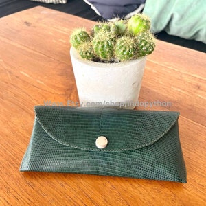 Op de afbeelding: Een donkergroene, getextureerde leren brillenkoker met een zilveren drukknoopsluiting. De koker is rechthoekig met een gebogen flap. Een kleine cactus in een pot staat op een houten tafel.