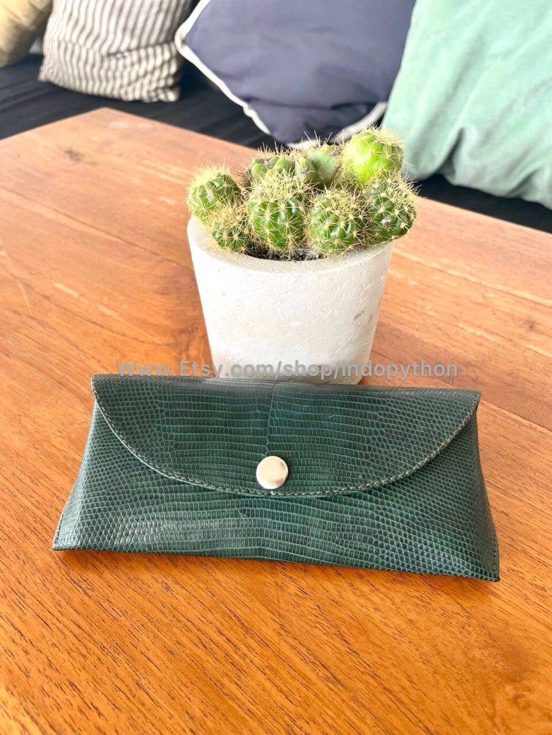 Op de afbeelding: Een donkergroene, getextureerde leren brillenkoker met een zilveren drukknoopsluiting. De koker is rechthoekig met een gebogen flap. Een kleine cactus in een pot staat op een houten tafel.
