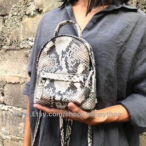 K&ouml;nnte beinhalten: Ein kleiner, wei&szlig;er und schwarzer Schlangenleder-Rucksack mit Rei&szlig;verschluss. Der Rucksack hat einen oberen Griff und verstellbare Tr&auml;ger.