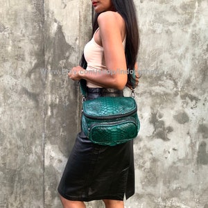 May include: A green snakeskin crossbody bag with a black strap and zipper accents. The bag is being worn by a person wearing a white tank top and a black skirt.