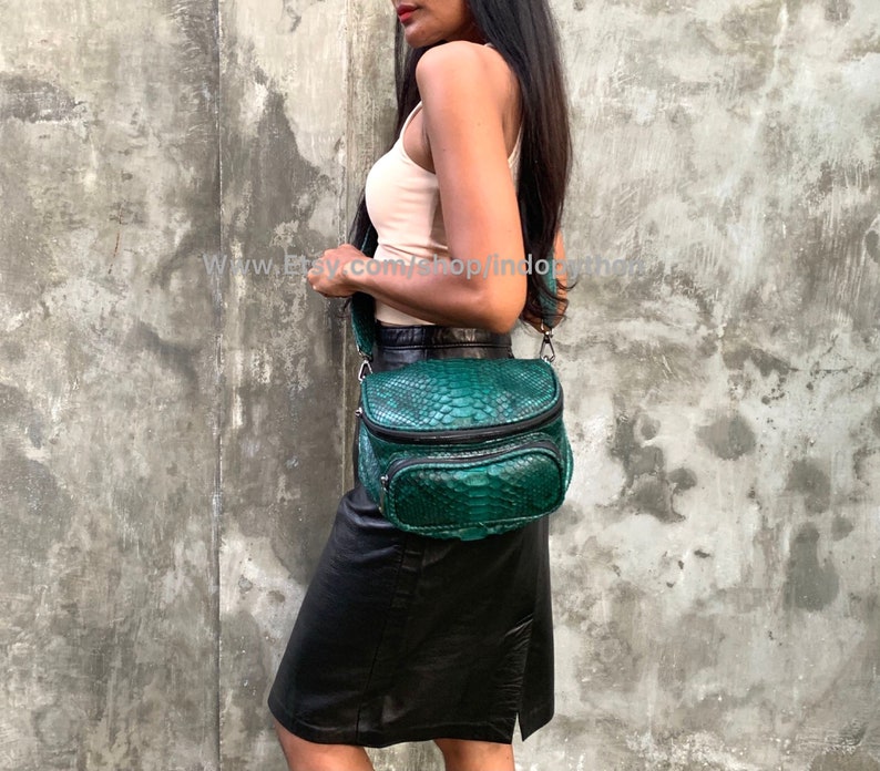 May include: A green snakeskin crossbody bag with a black strap and zipper accents. The bag is being worn by a person wearing a white tank top and a black skirt.