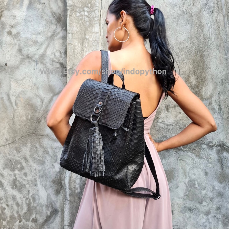 May include: Black leather backpack with a snakeskin pattern and a tassel. The backpack has a flap closure and a zipped pocket on the front.
