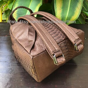 May include: A brown backpack with a snakeskin pattern. The bag has adjustable straps and silver-colored metal hardware. The top of the bag has a handle. The bag is sitting on a dark wooden surface.