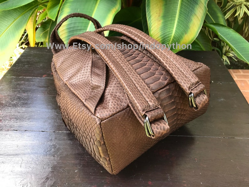 May include: A brown backpack with a snakeskin pattern. The bag has adjustable straps and silver-colored metal hardware. The top of the bag has a handle. The bag is sitting on a dark wooden surface.