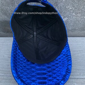 May include: A blue baseball cap with a snakeskin pattern. The inside of the cap is black with a white stitching detail. The cap has a black adjustable strap with a silver buckle. The text "www.Etsy.com/shop/indopython" is printed on the inside of the cap.